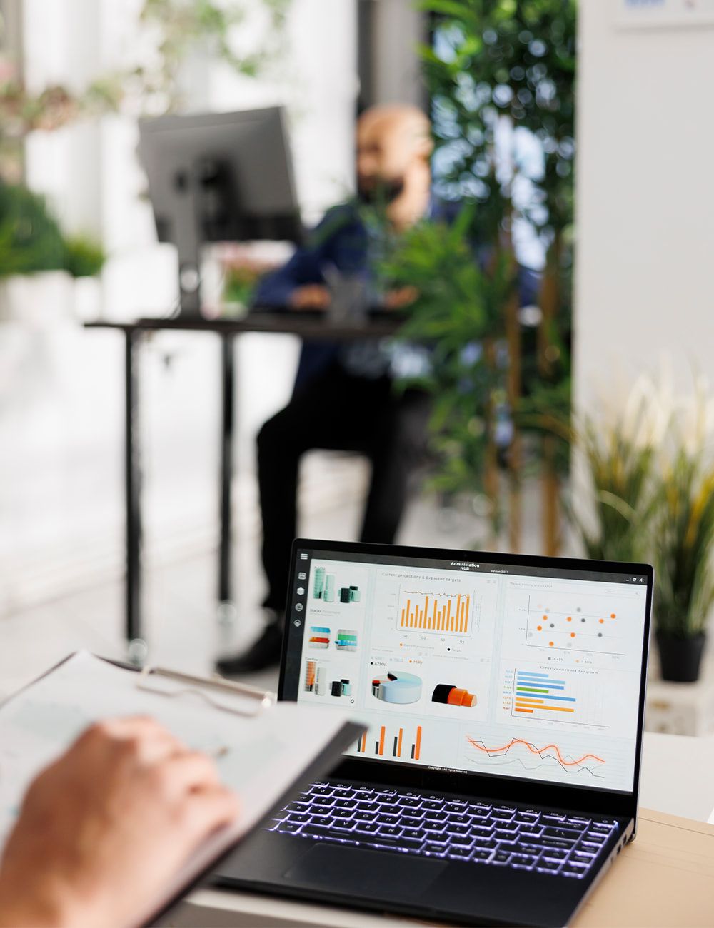Ordinateur portable ouvert sur un tableau de bord de suivi, une main annote un document imprimé au premier plan