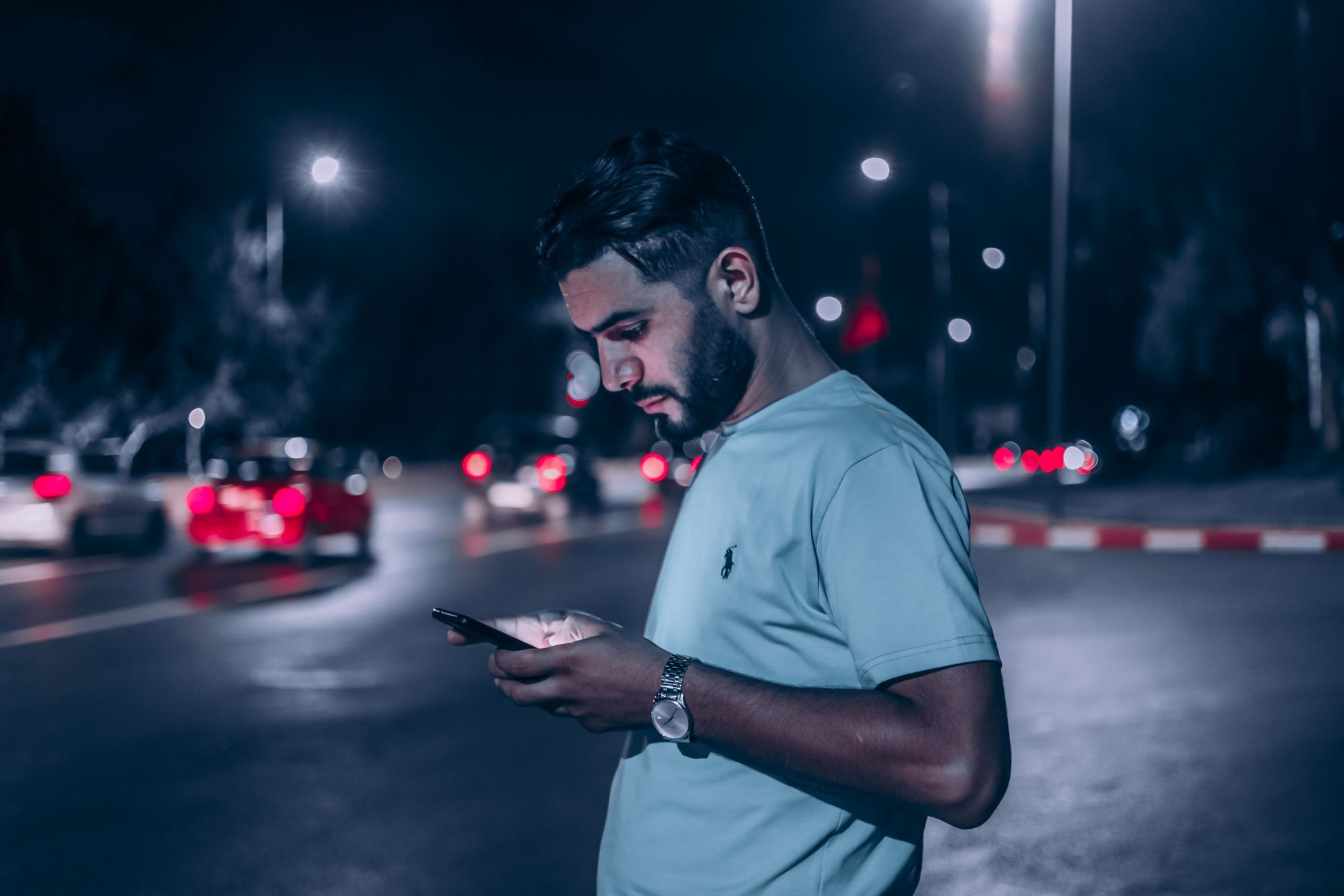 Un homme lit son téléphone la nuit au bord d'une route, les feux arrière des voitures flous derrière lui