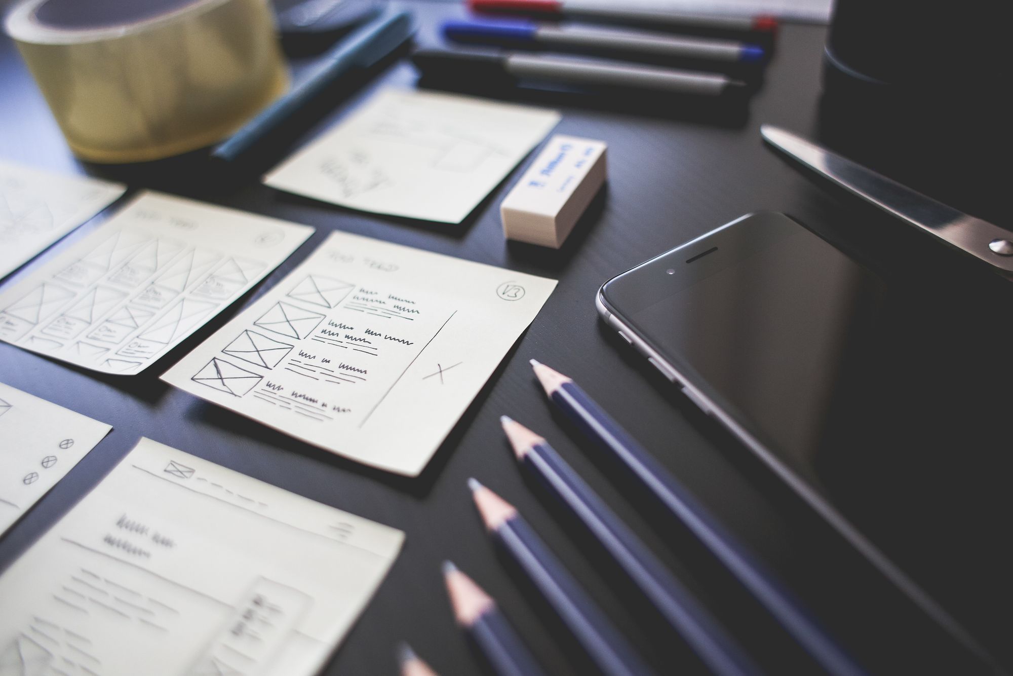 Plusieurs fiches de maquette d'interface dessinées à la main, posées sur un bureau sombre avec des crayons et un téléphone
